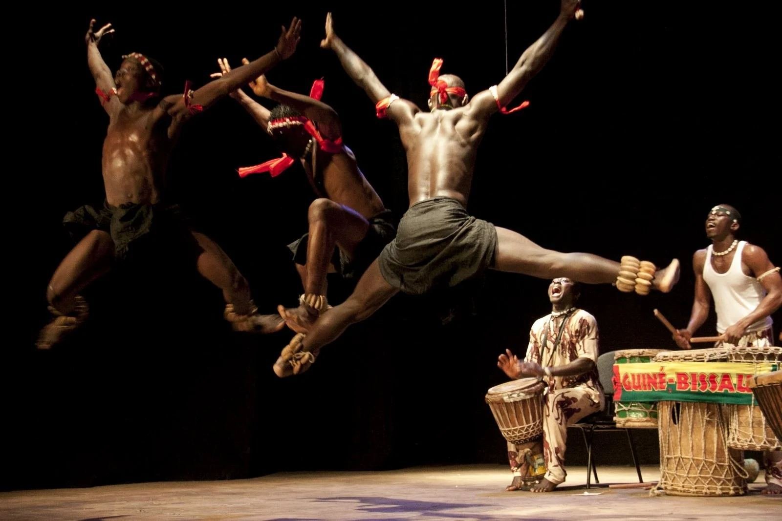 Bailarinos dos Netos de Bandim em salto, com percussionistas ao fundo