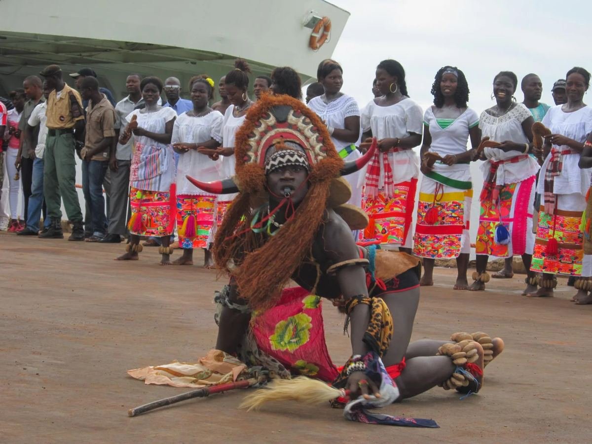 Bailarino mascarado num desfile, com o grupo de mulheres em fila atrás