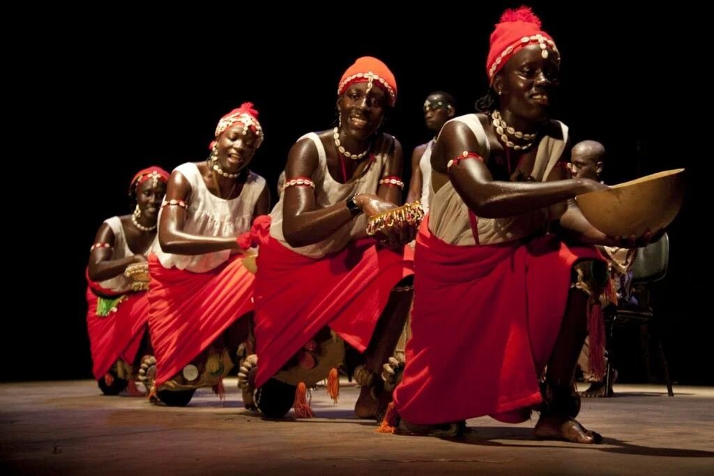 Bailarinas de vermelho ajoelhadas em palco, com cabaças